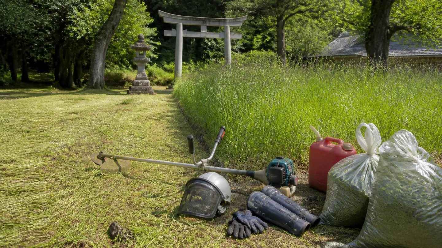 草を刈った範囲と未作業の範囲の境に、草刈機、防護面、すね当て、手袋、燃料容器、刈草袋を置いた架空の神社境内