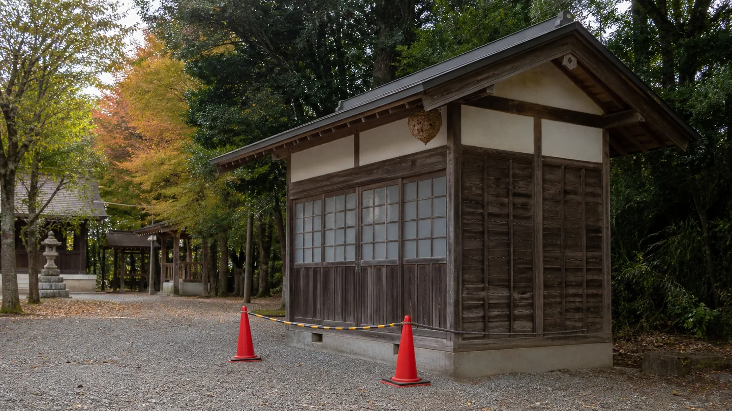 架空の小さな神社境内で、蜂の巣が木造建物の高い軒下にあり、無人の砂利道を2本の赤いコーンとロープで立入防止している秋の安全点検場面