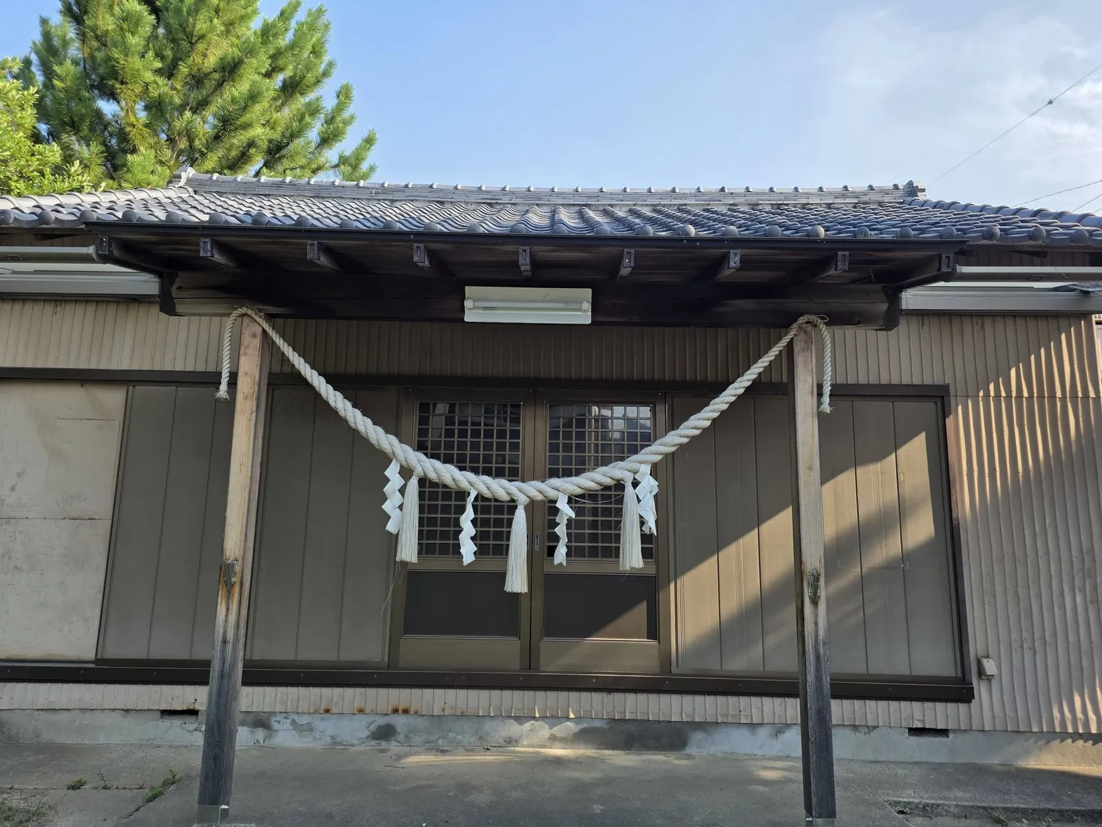 社殿正面の近景。二本の柱に注連縄を渡し、正面は格子入りの引戸とする。軒下に照明を吊る。