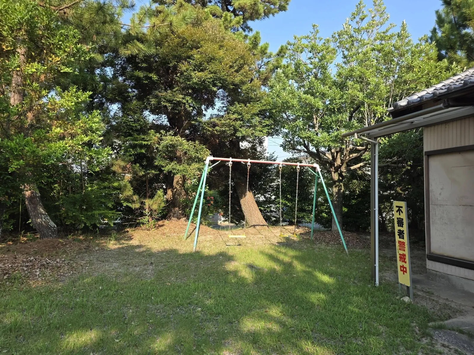 境内の社叢。マツと常緑広葉樹の大木が茂り、その根元にブランコが据えられている。右端は社殿。