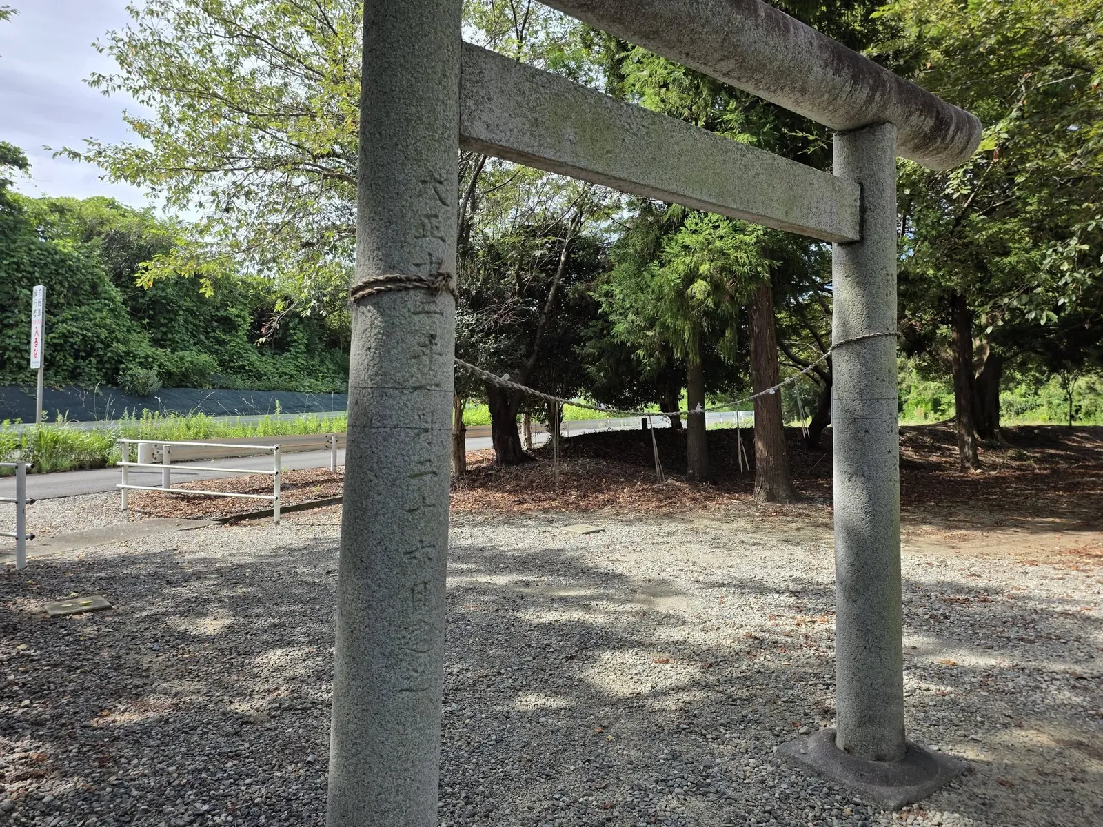 鳥居の柱に刻まれた年号銘。奥に注連縄を渡した参道と社叢、道路のガードレールが見える。