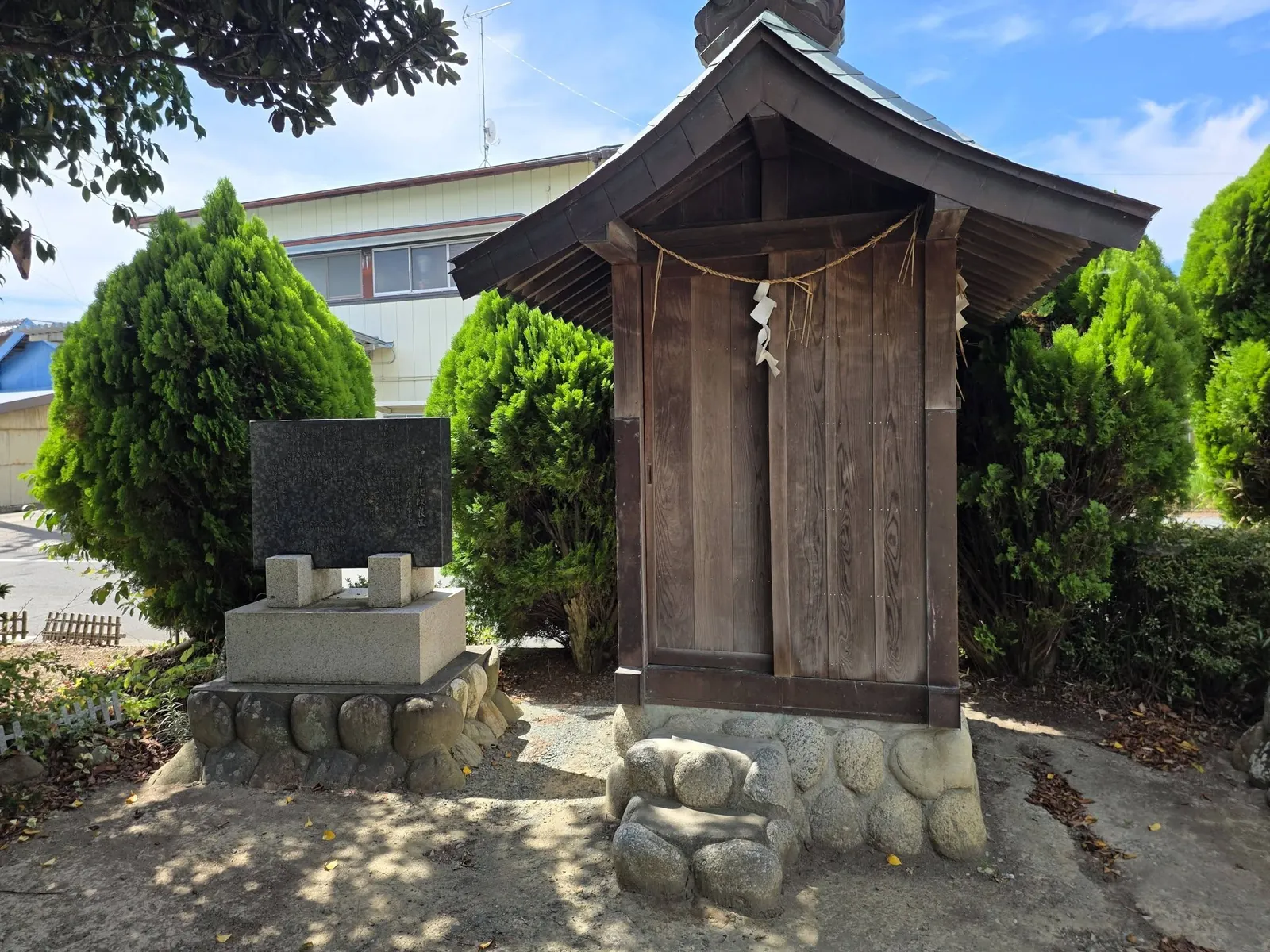 小祠の正面。しめ縄と紙垂が掛かる木製の扉と、脇に立つ石碑。