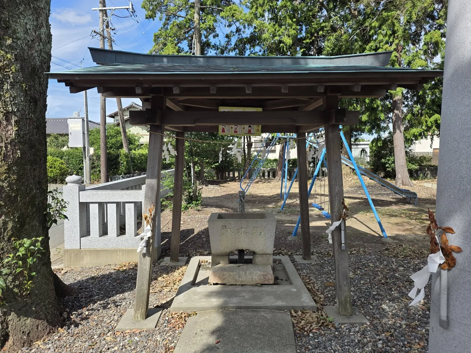 屋根付きの手水舎。「浄水鉢」と刻まれた水盤があり、奥に子供用遊具と住宅が見える。